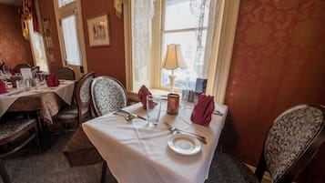 A dining area with a table set for two, featuring a white tablecloth, two chairs with floral upholstery, a candle, and a window with lace curtains.