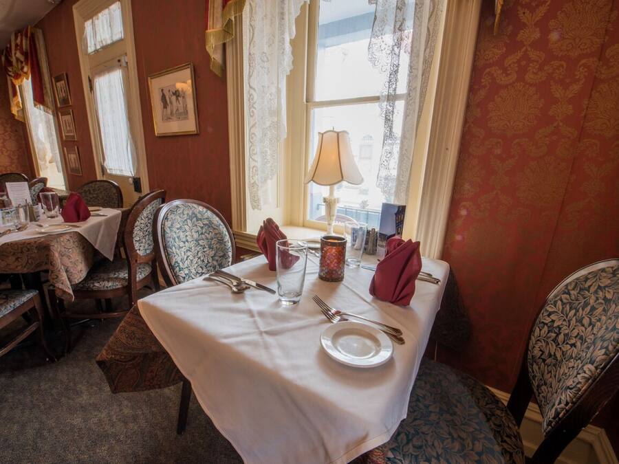 A dining area with a table set for two, featuring a white tablecloth, two chairs with floral upholstery, a candle, and a window with lace curtains.