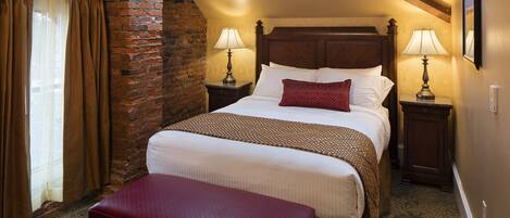 Una habitación de hotel con una cama grande, un banco, dos mesitas de noche con lámparas, una pared de ladrillo y una ventana con cortinas.