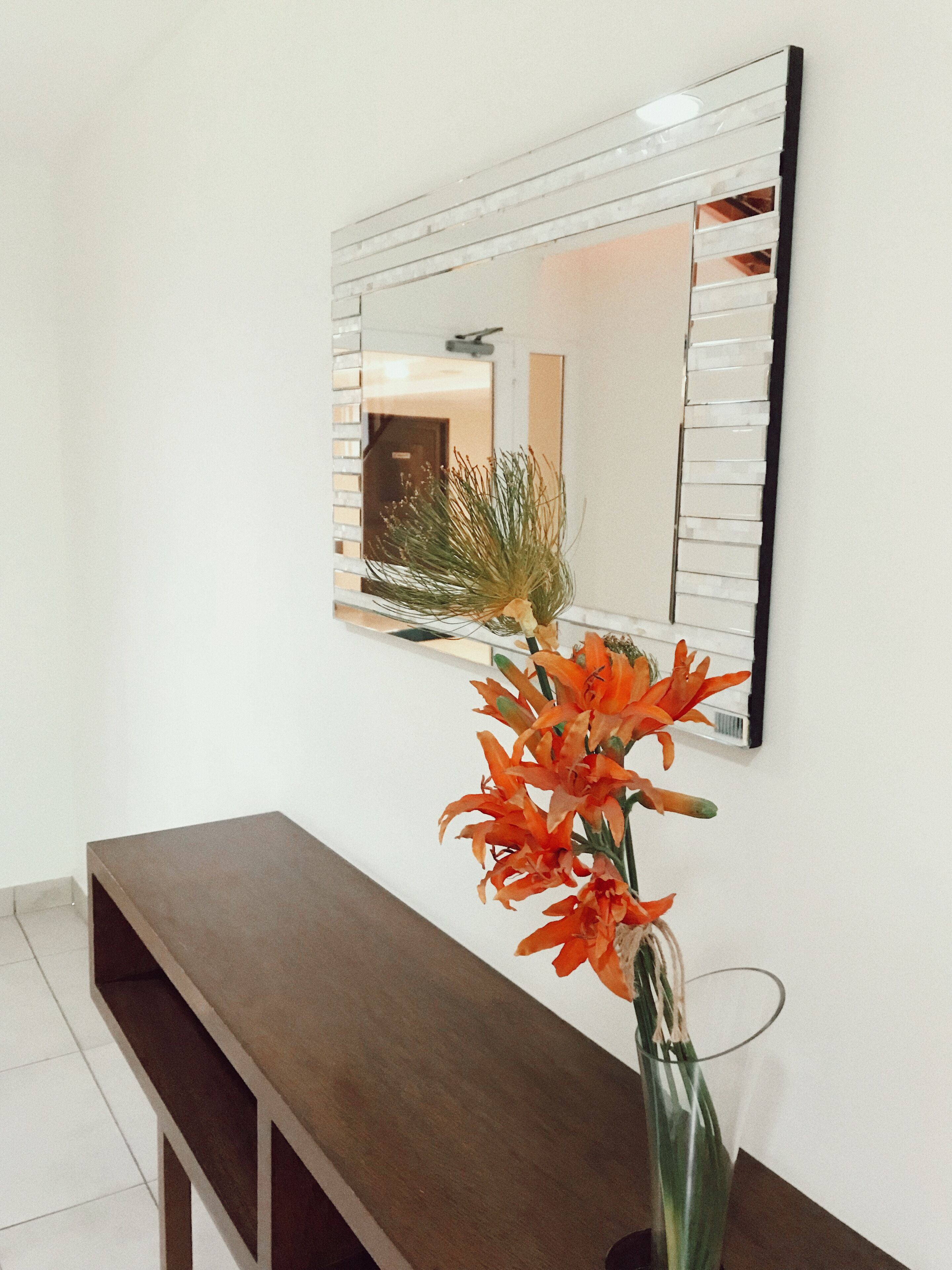 Ein modernes Interieur mit einem Holzkonsolentisch, einem Spiegel mit geometrischer Rahmen und einer Vase mit orangefarbenen Blumen.