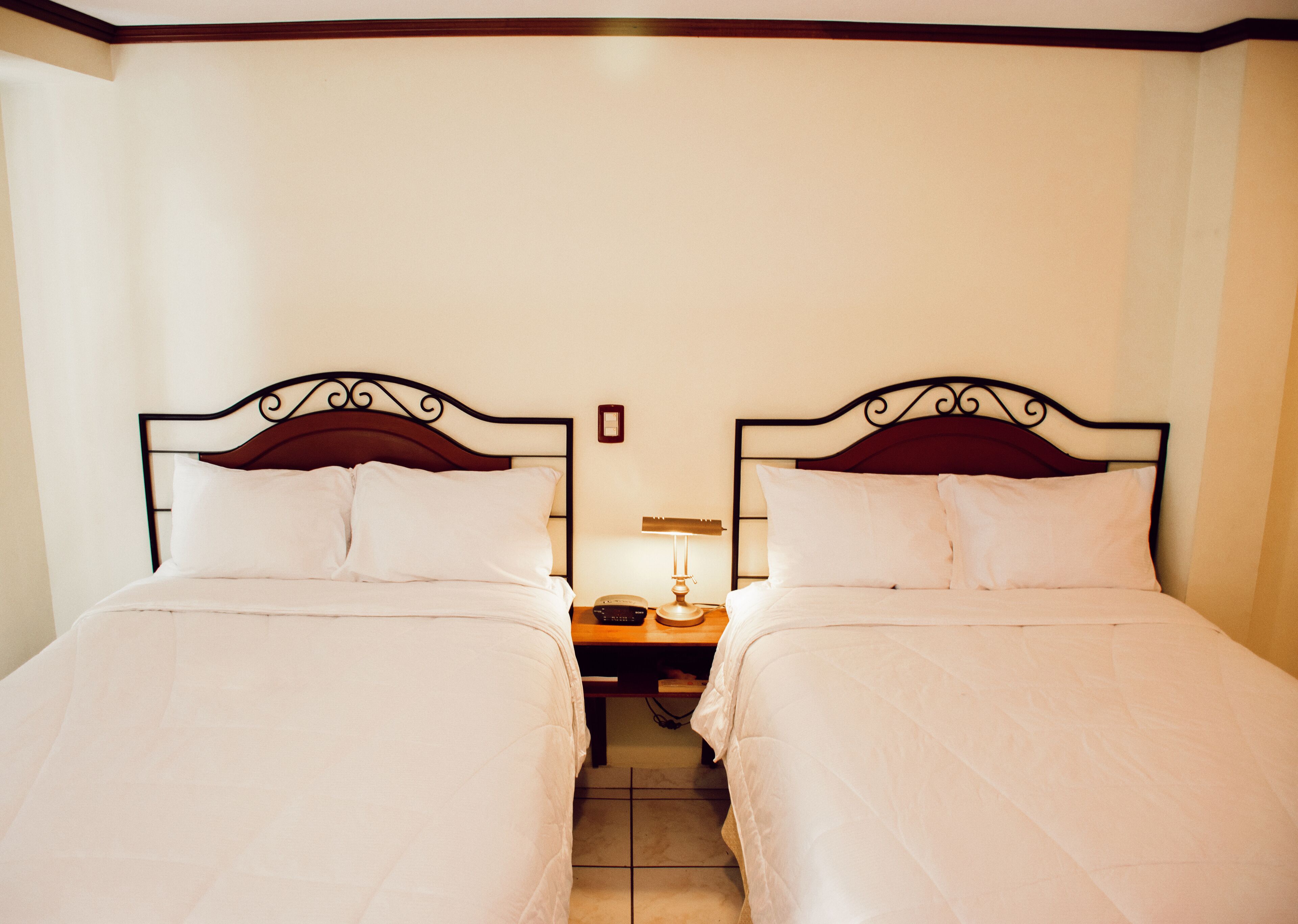 A hotel room with two single beds, a nightstand with a lamp, and a small table between the beds.