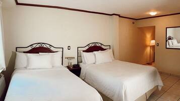 A hotel room with two single beds, a nightstand with a lamp, a wall-mounted mirror, and a tiled floor.