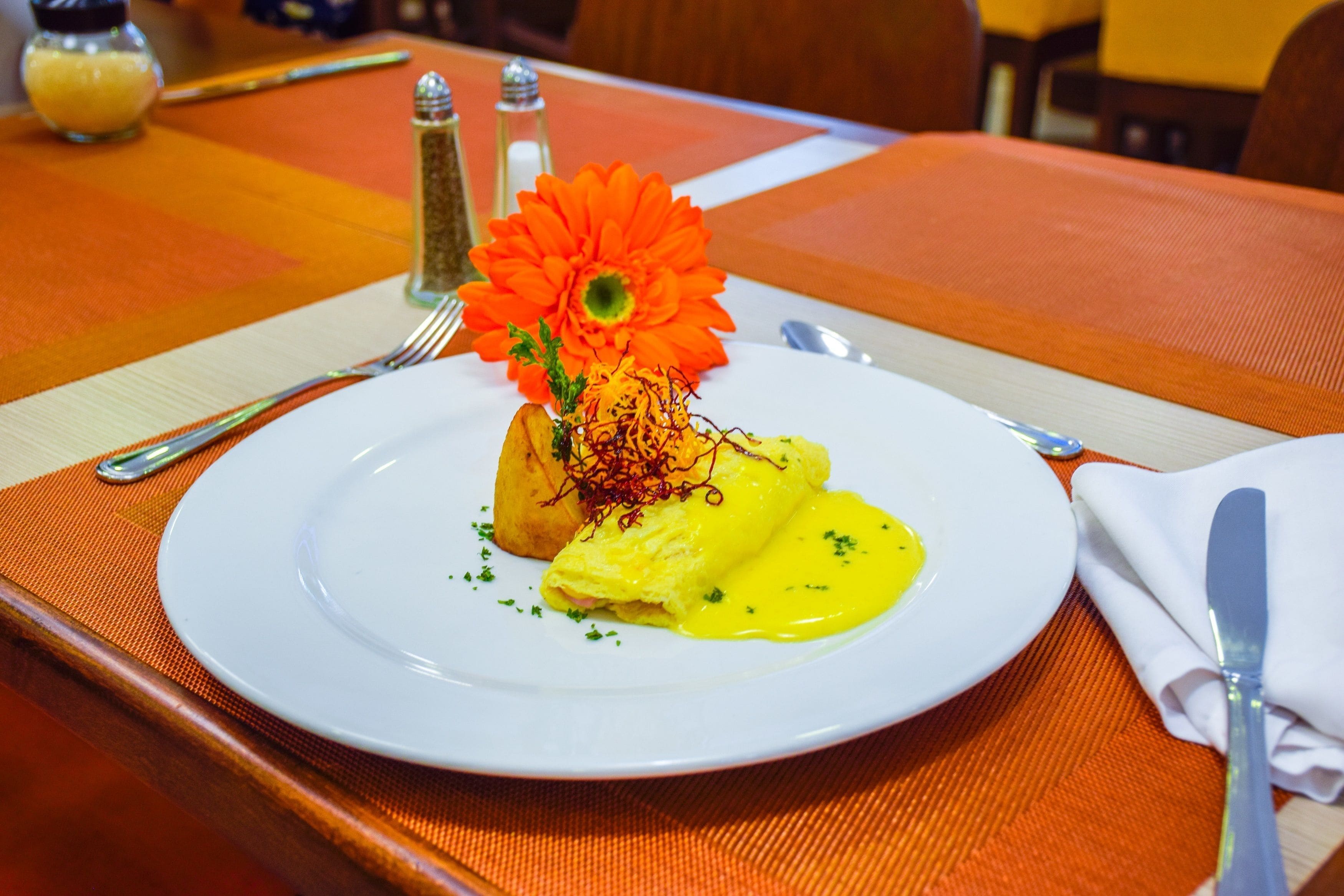 Um prato branco com molho amarelo, uma flor e um acompanhamento sobre uma toalha laranja.