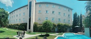 Un hotel de varios pisos con piscina, mobiliario exterior y abundante vegetación.