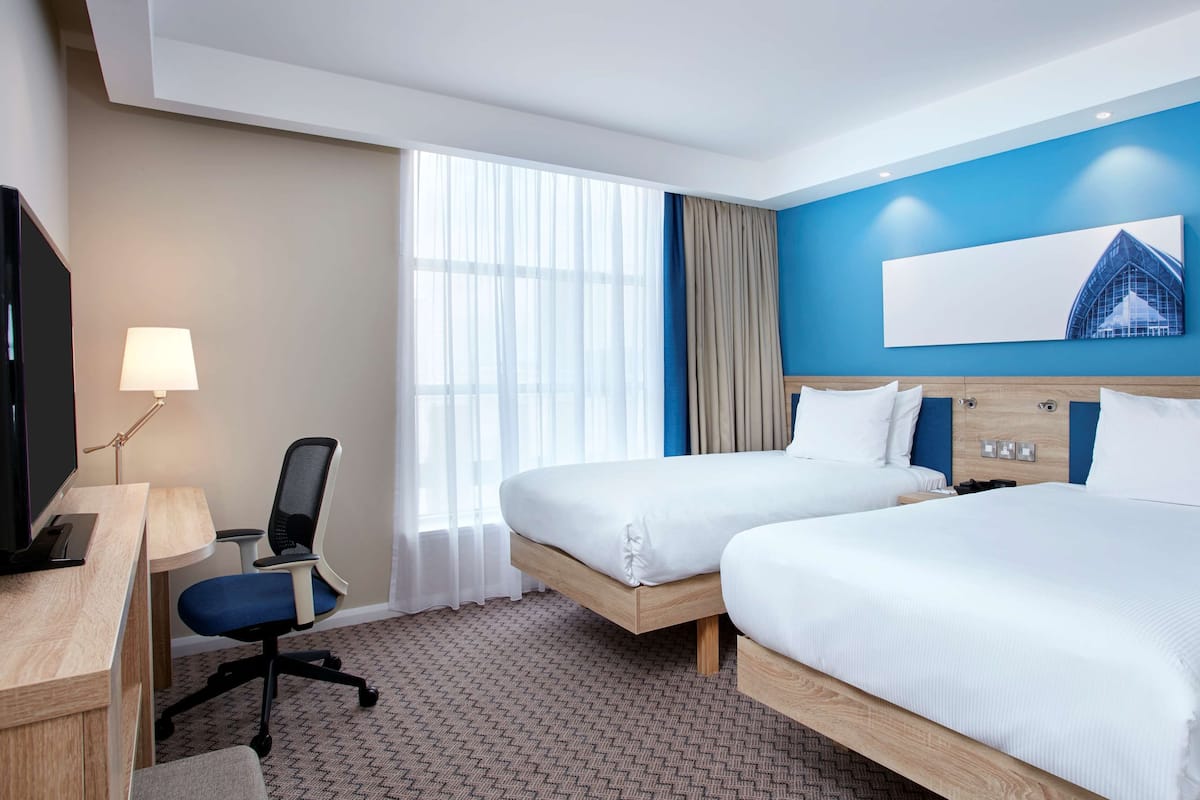 A hotel room with two beds, a desk with a chair, a TV, and a large window with curtains.