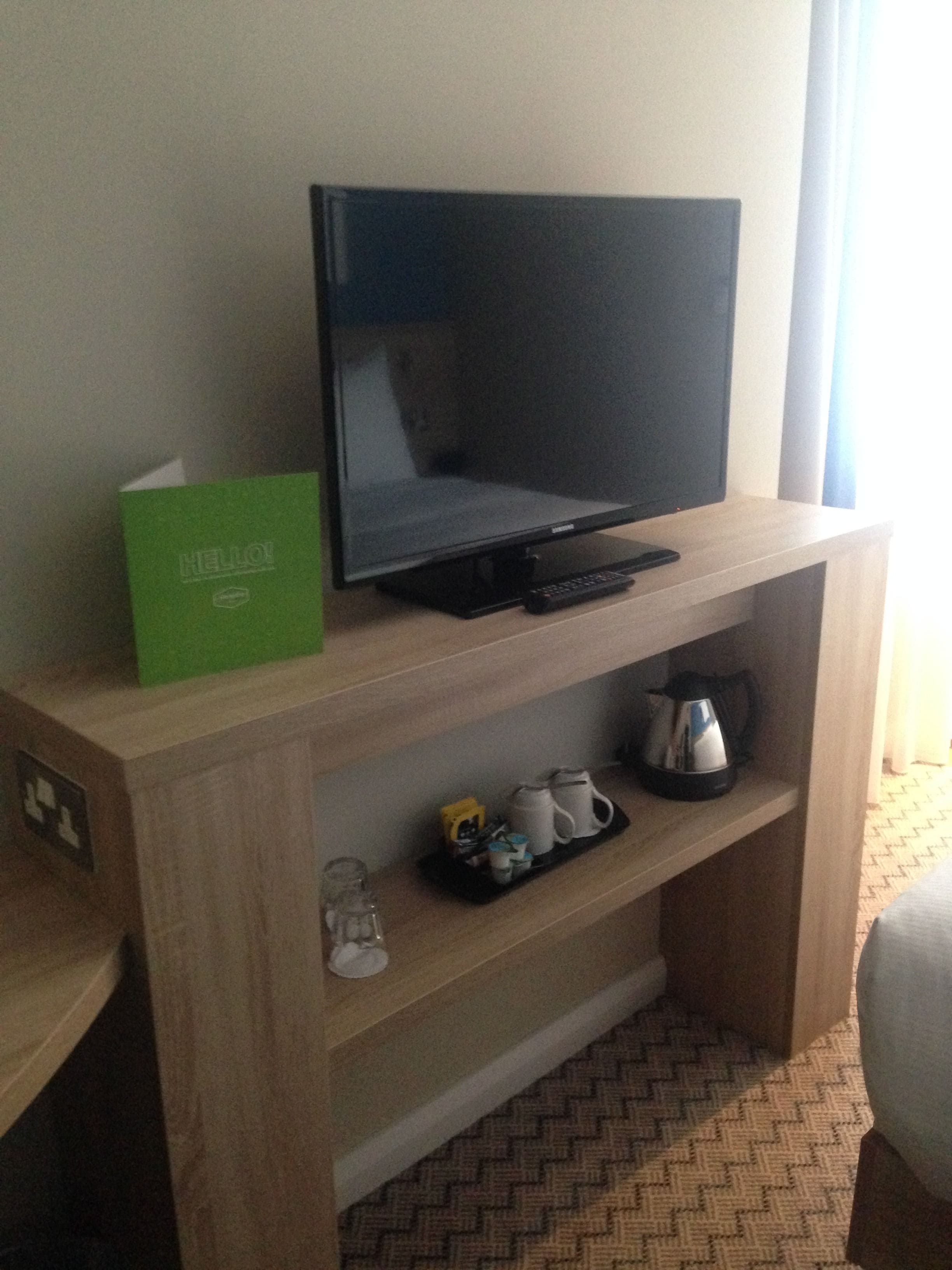 A hotel room with a flat-screen TV on a wooden stand, a green "HELLO!" sign, a tray with mugs and a kettle, and a patterned carpet.