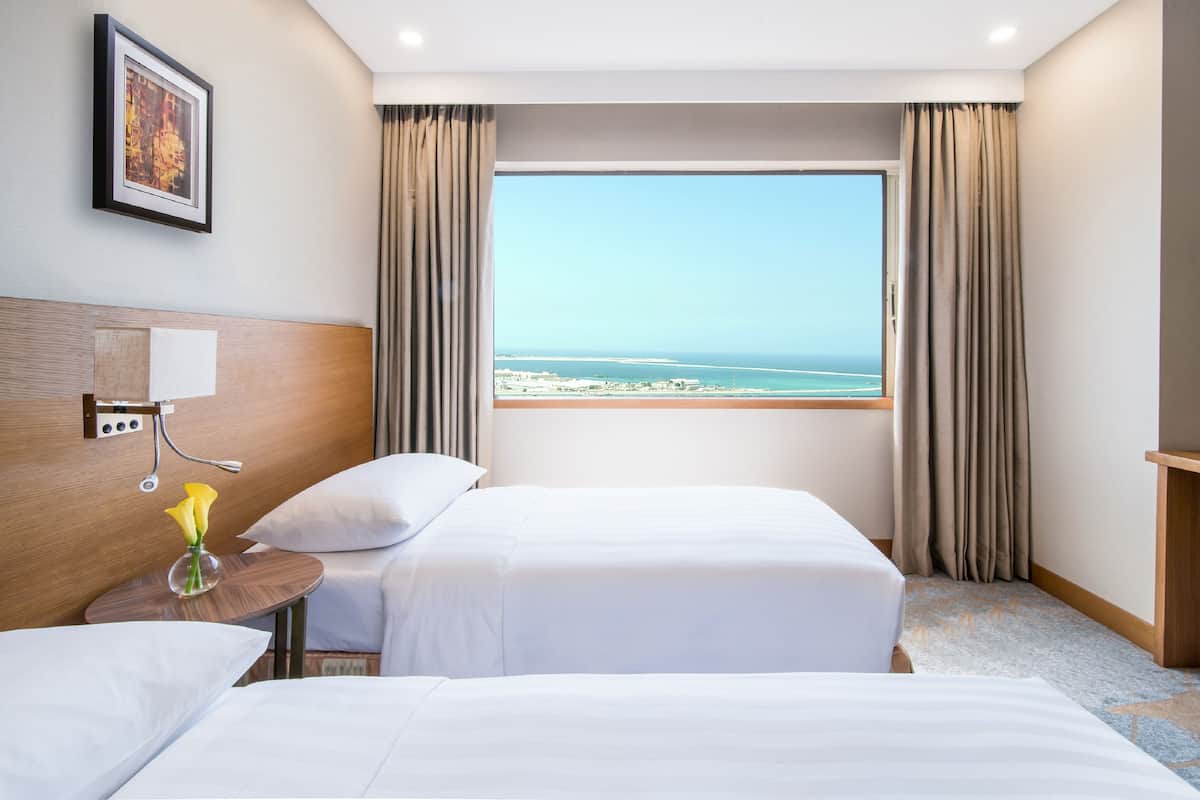 Habitación de hotel con dos camas, un ventanal con vista a la playa, un cuadro enmarcado en la pared y una mesita con un jarrón de flores.