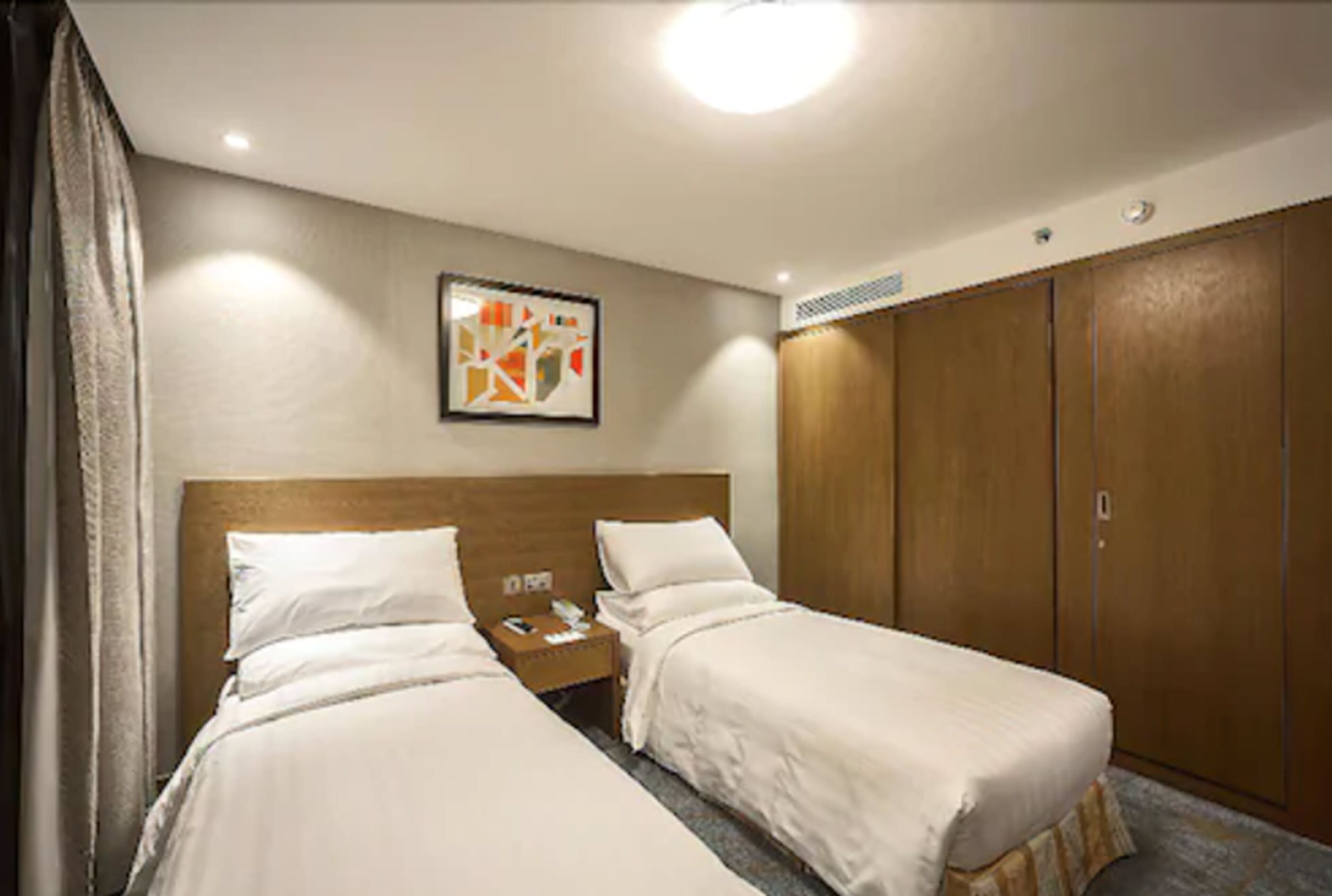 A hotel room with two single beds, a wooden wardrobe, a nightstand with a lamp, and a framed abstract artwork on the wall.