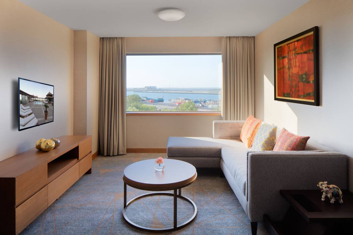Una habitación de hotel moderna con un ventanal grande, un sofá, una mesita de centro redonda y un televisor sobre un mueble de madera.