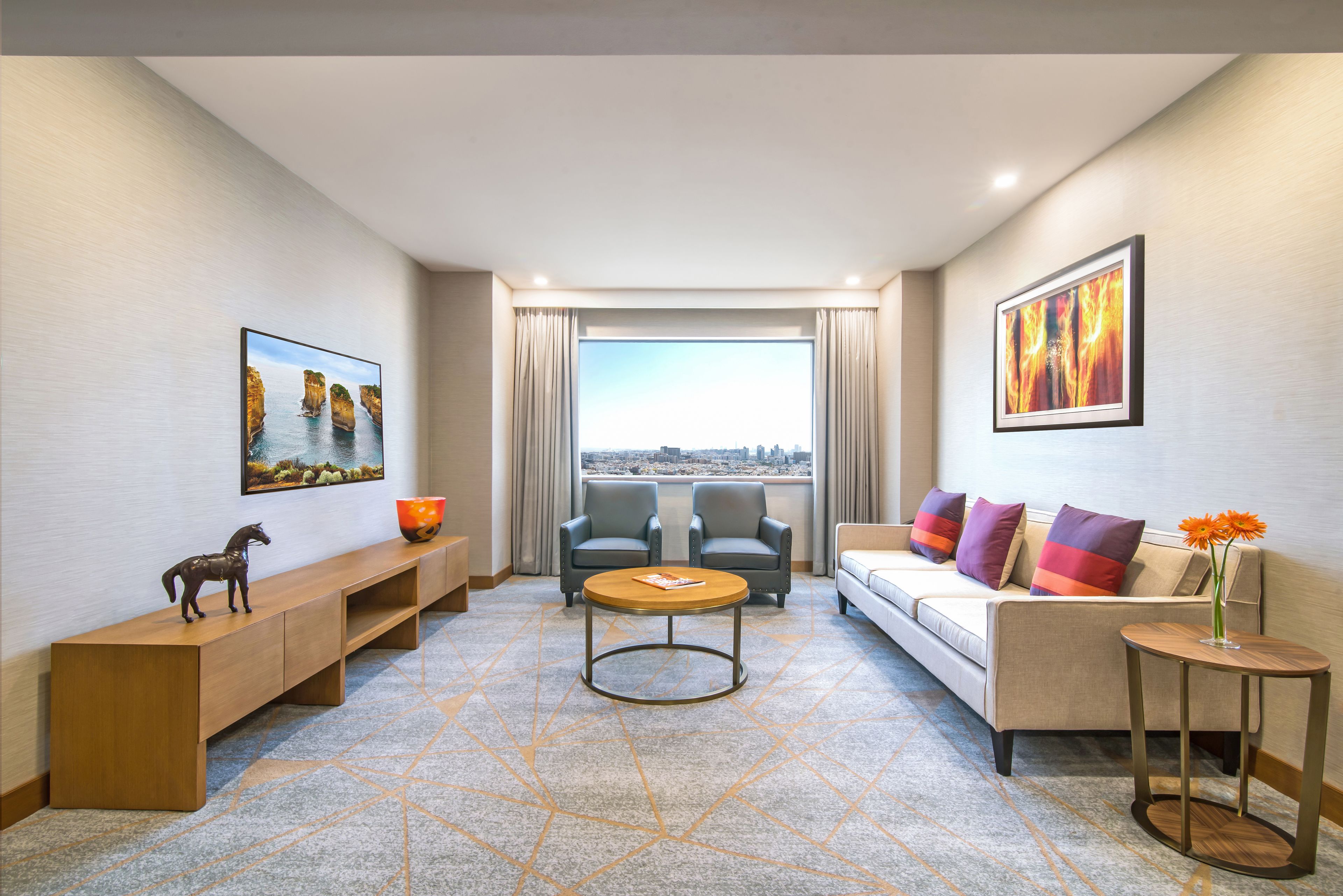 A modern hotel room with a sofa, two armchairs, a coffee table, a console table with a horse figurine, and artwork on the walls.
