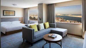 Hotelkamer met een bed, een grijze bank met gele kussens, een ronde salontafel met een tijdschrift en een groot raam met uitzicht op de stad en de haven.