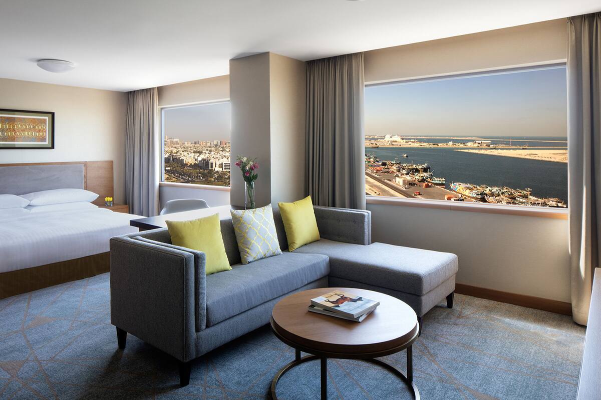 Habitación de hotel con cama, sofá gris con cojines amarillos, una mesita de centro redonda con una revista y un amplio ventanal con vistas a la ciudad y al puerto.