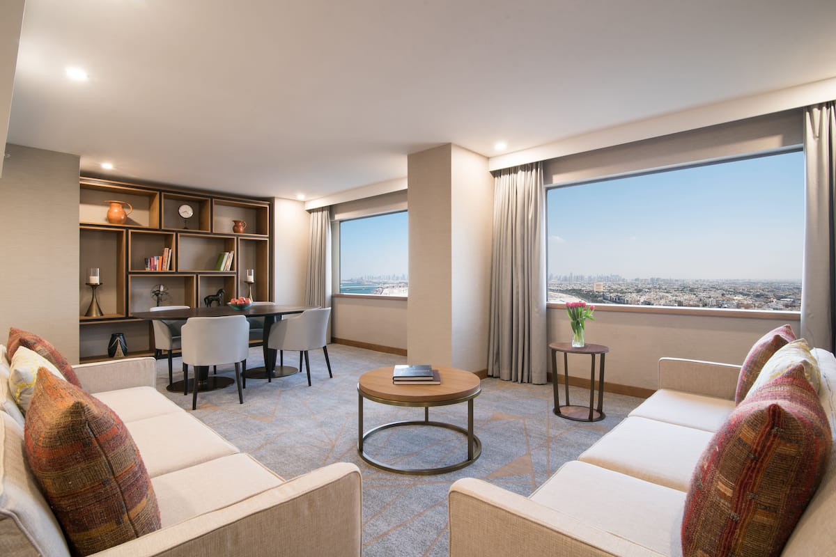Una habitación de hotel moderna con un sofá, una mesa redonda, un rincón de comedor con sillas, una estantería y un amplio ventanal con vistas a la ciudad.