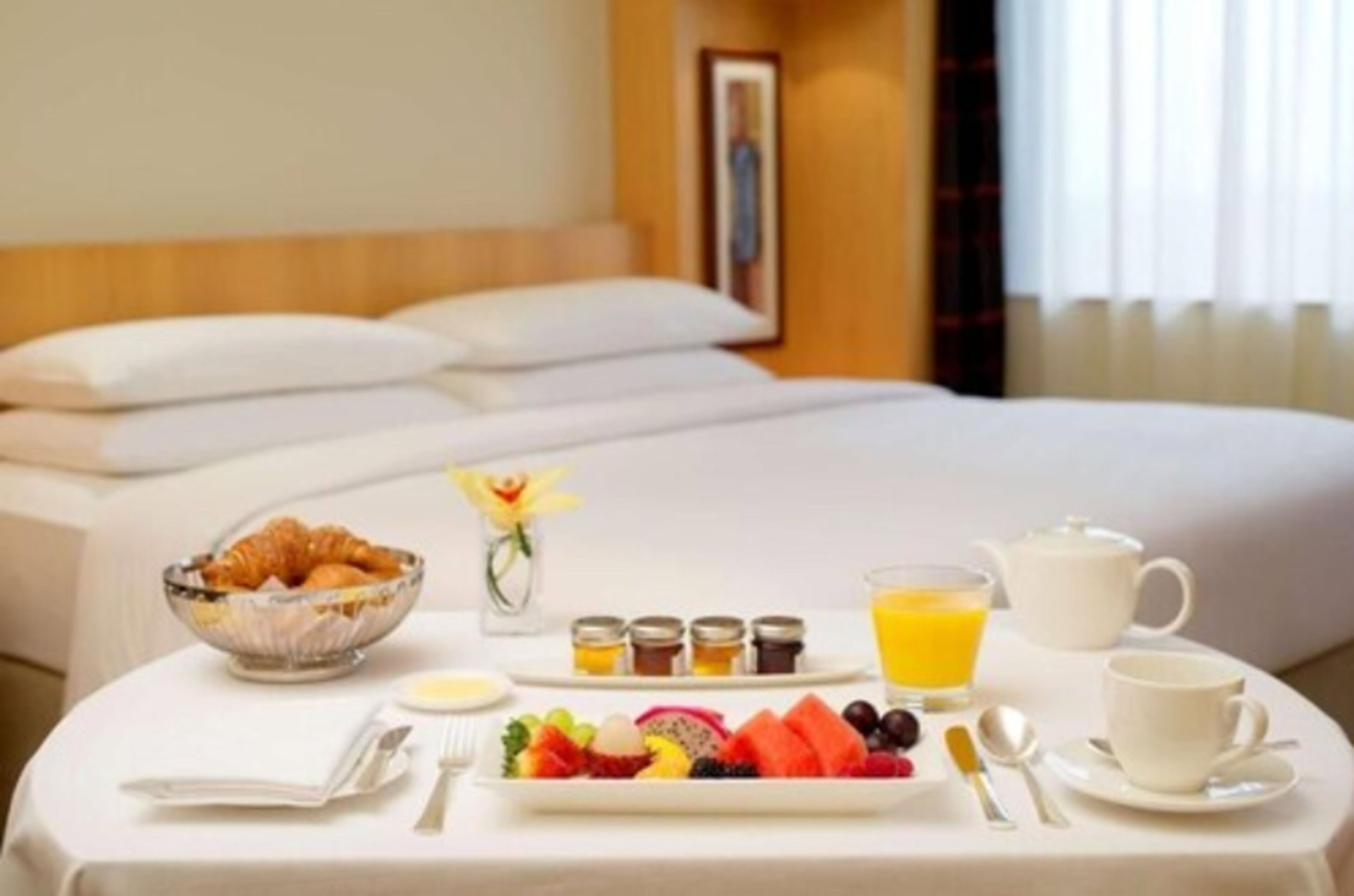 Un lit parfaitement fait, avec du linge de lit blanc, un plateau de petit-déjeuner comprenant des croissants, des fruits et des boissons, et un vase contenant une seule fleur jaune.