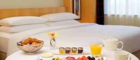 Un letto rifatto con lenzuola bianche, un vassoio per la colazione con croissant, frutta e bevande, e un vaso con un singolo fiore giallo.