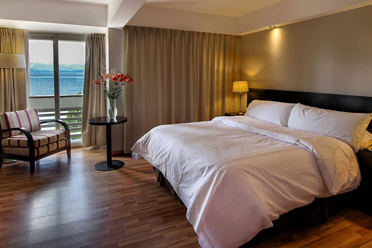 A hotel room with a large bed, a chair, a small table with a vase, and a view of the sea through a large window.
