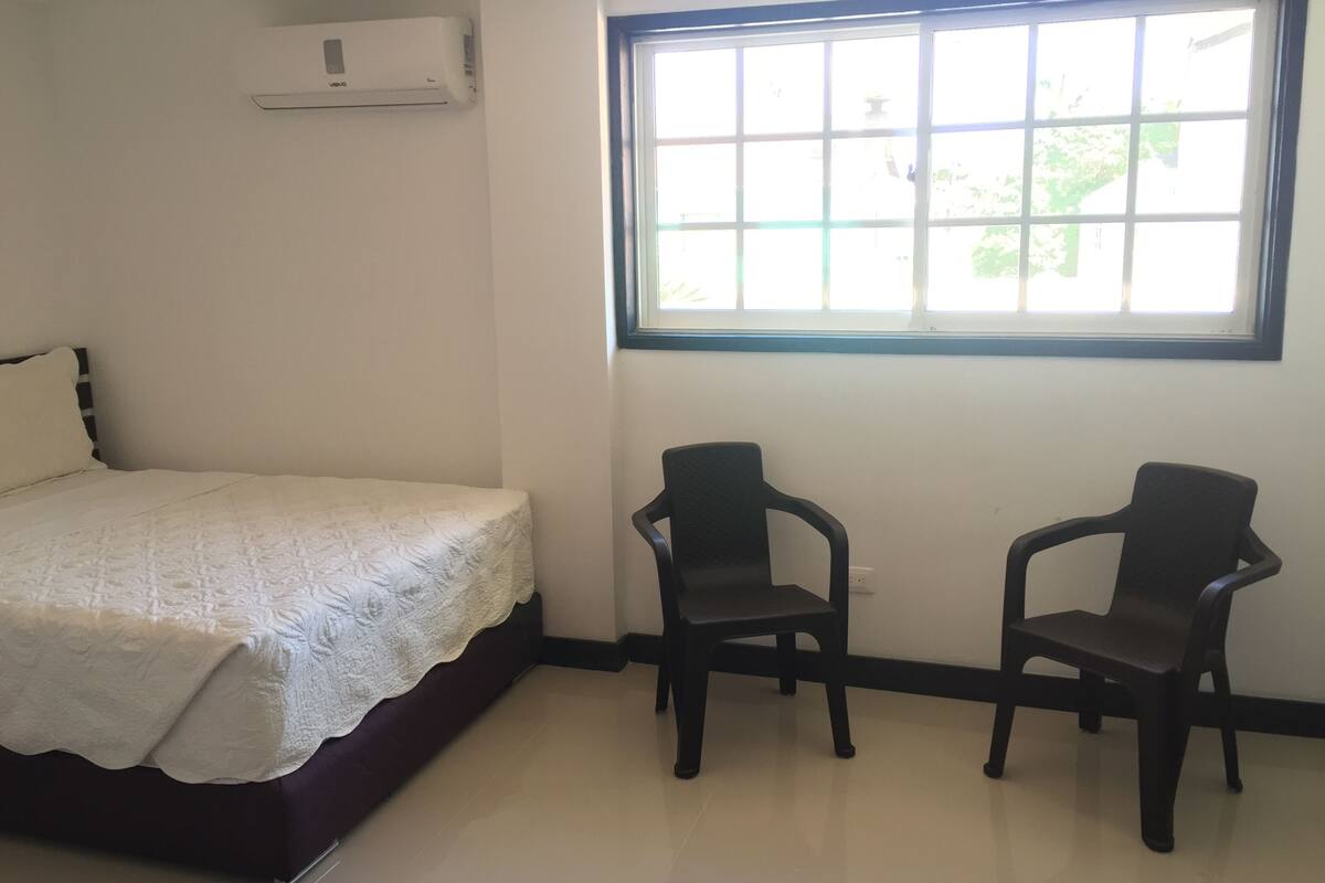 A bedroom with a bed, two chairs, a window, and an air conditioning unit.