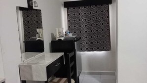 A small, clean room with a bed, a desk, and a chair. There is a window with patterned curtains, an air conditioning unit, and a blue plastic bag on the floor.