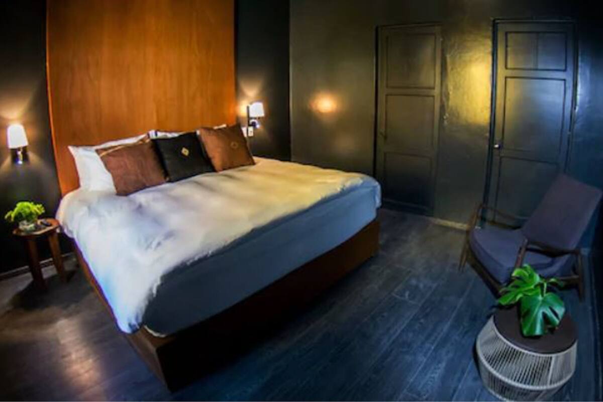 A modern hotel room with a large bed, wooden headboard, two bedside lamps, a small round side table with a plant, and a chair.