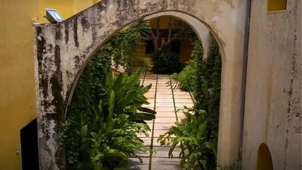 An arched passageway with a tiled floor, surrounded by greenery and a yellow wall.