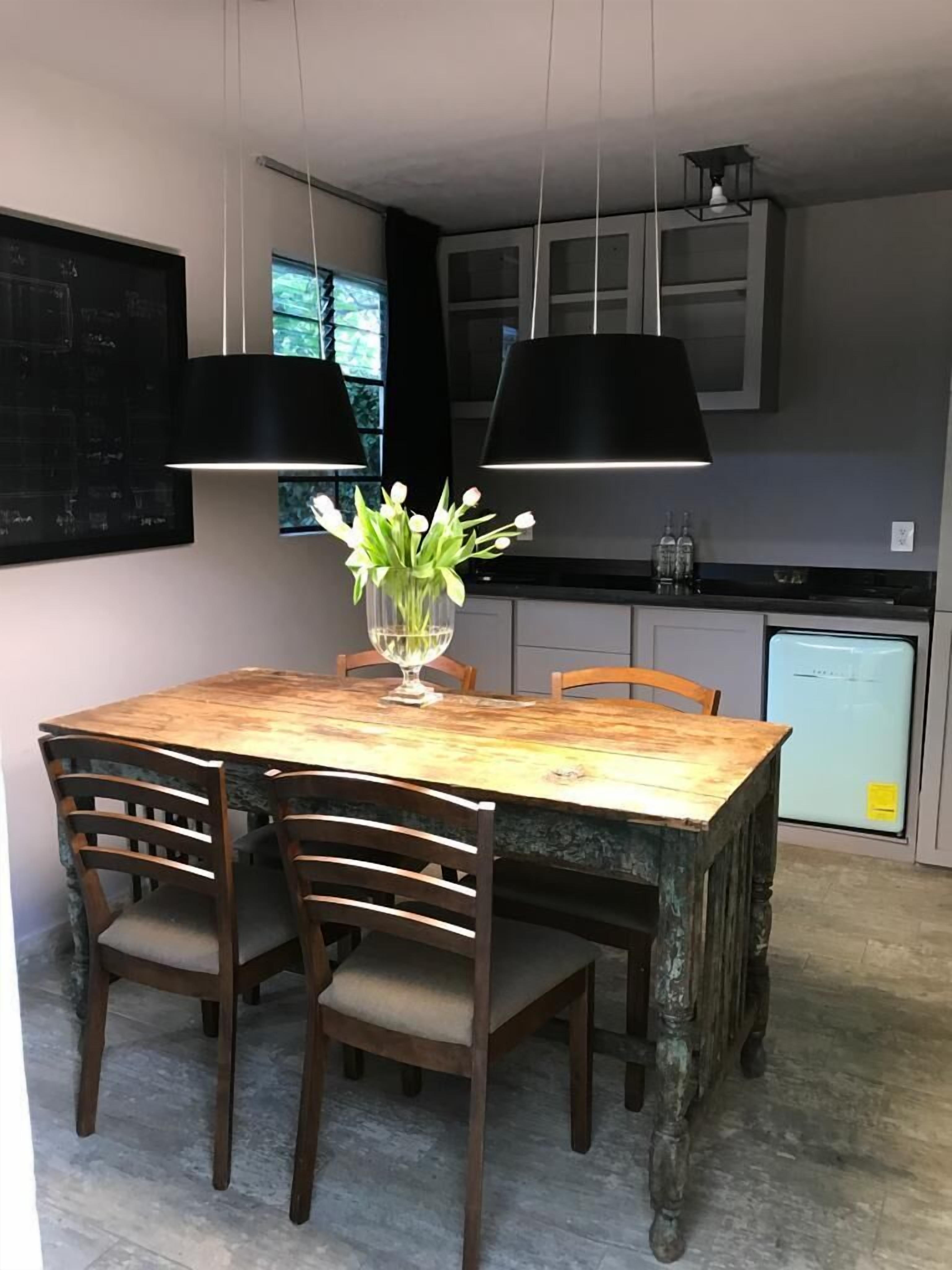 Un comedor con una mesa de madera, cuatro sillas, un jarrón con flores, lámparas colgantes, un refrigerador y armarios.