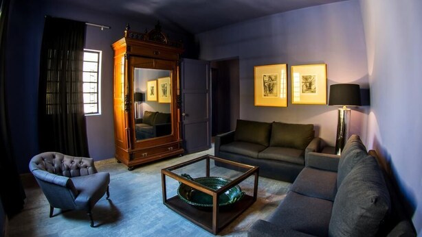 Uma sala de estar com parede azul escura, um armário de madeira com quadros emoldurados, um sofá cinza, uma mesa de centro de vidro e um abajur.