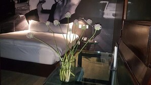 A bedroom with a bed, a glass coffee table with a vase of flowers, and a poster on the wall.