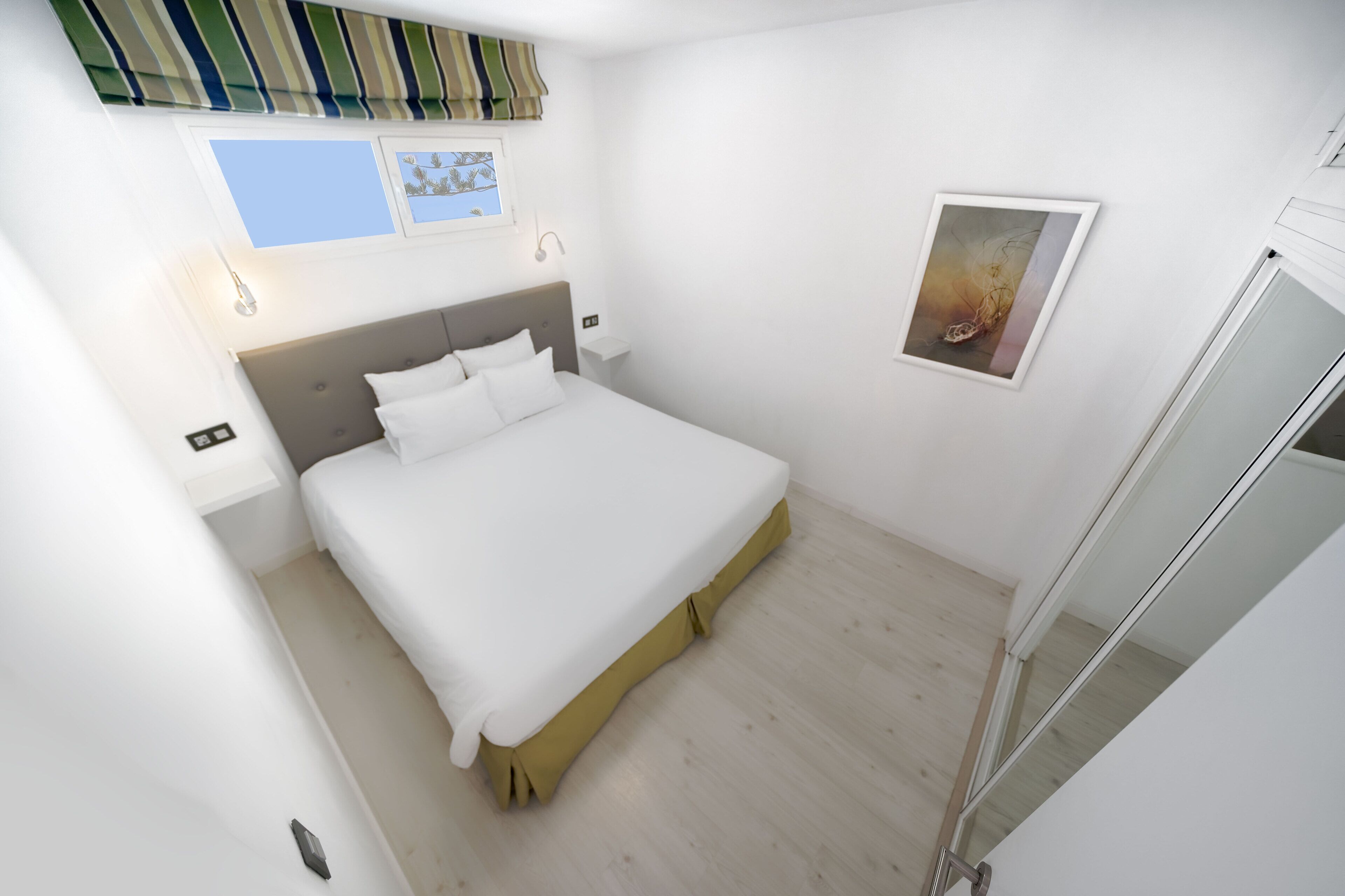 Una habitación de hotel moderna con una cama grande, una mesita de noche, un ventanal con vistas y un cuadro en la pared.