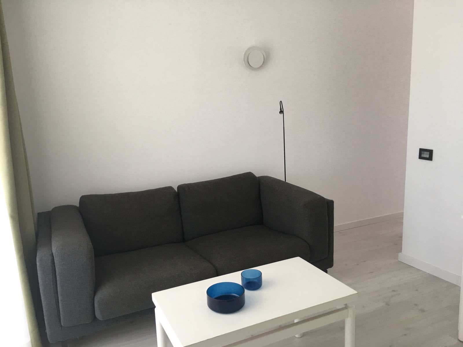 Un salon moderne avec un canapé gris, une table basse blanche ornée d’objets bleus et un mur blanc sobre agrémenté d’un détecteur de fumée.
