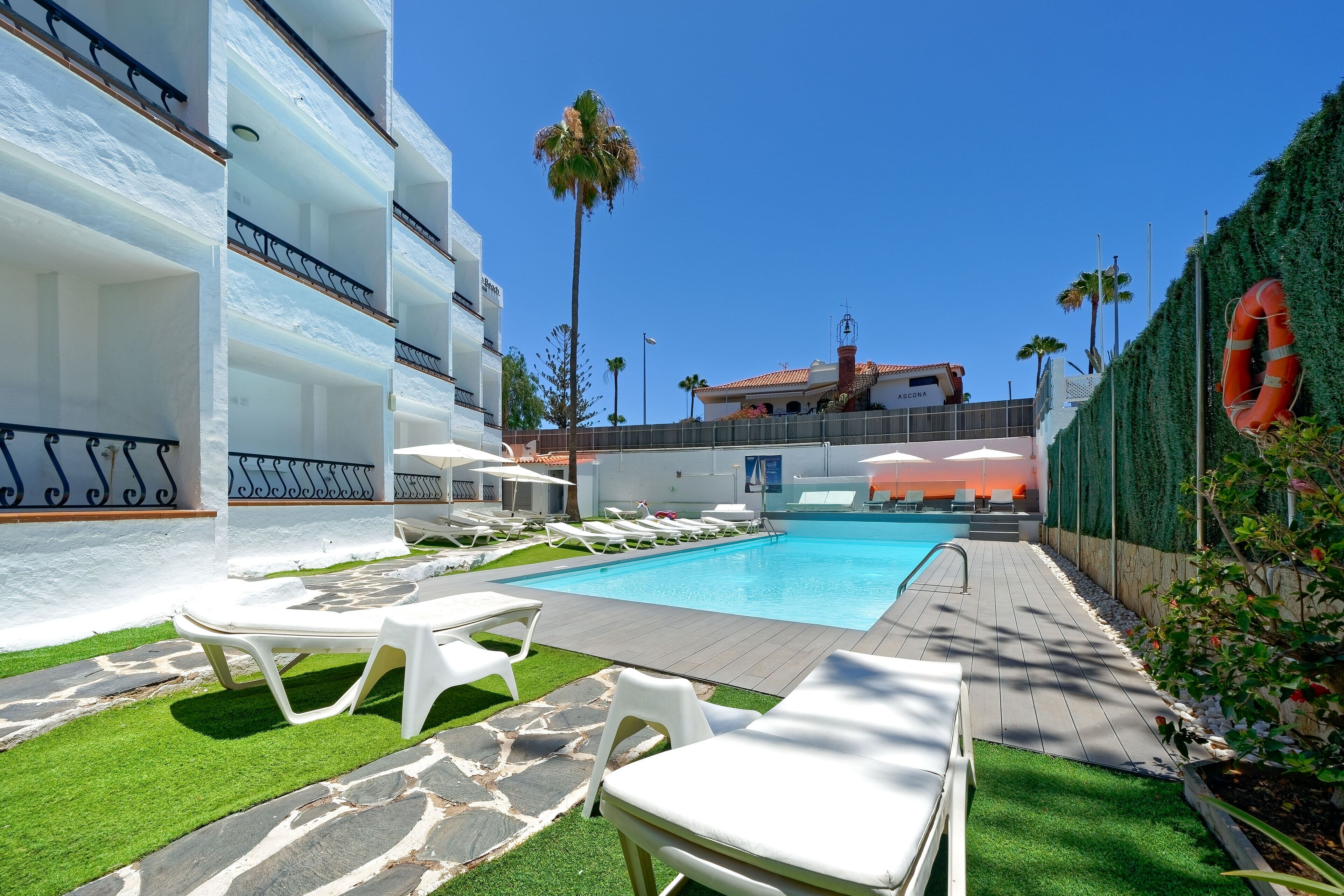 A hotel pool area with sun loungers, a swimming pool, and a view of the surrounding buildings.