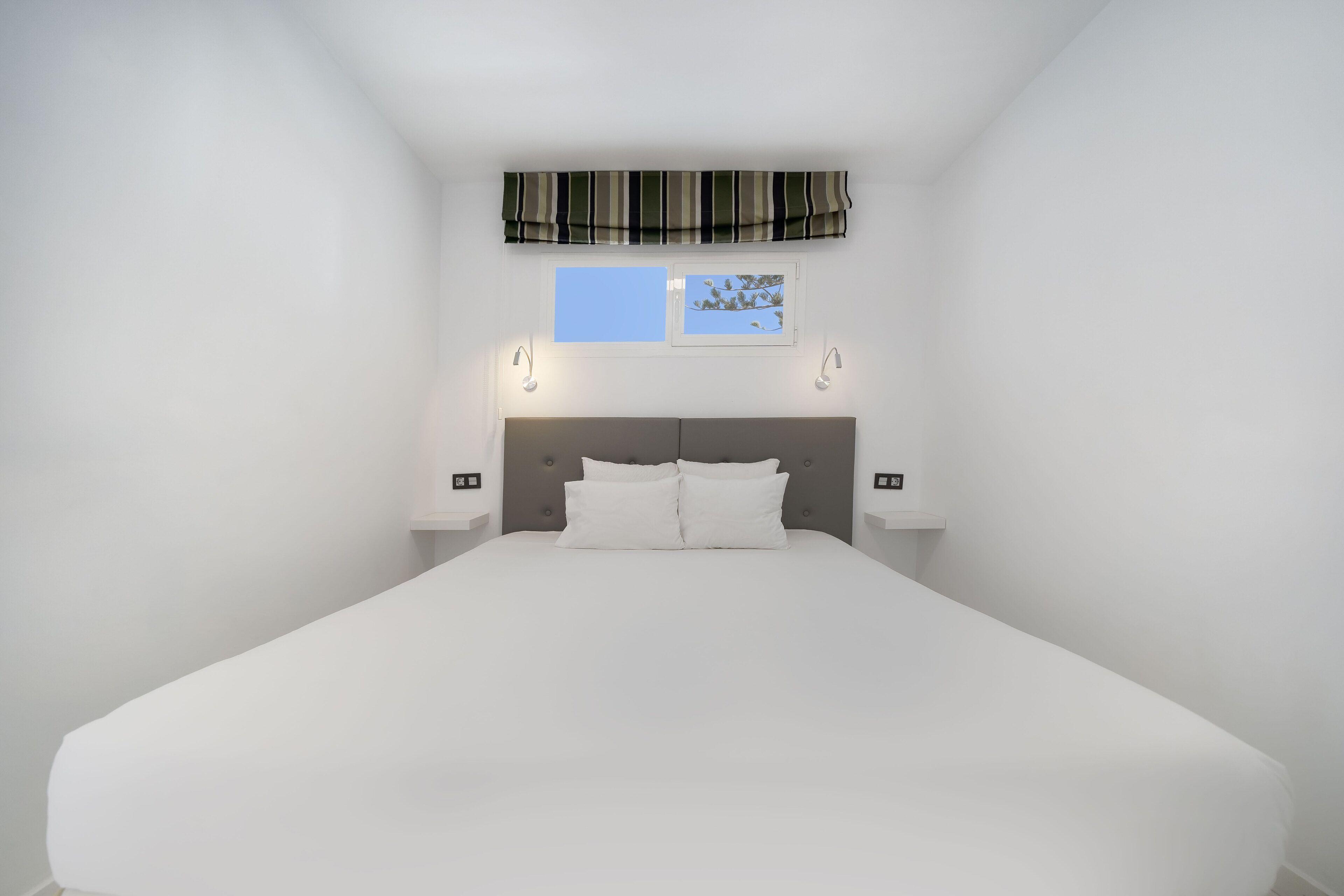 A neatly made bed in a room with a striped valance above a window, two bedside lamps, and a small picture frame.