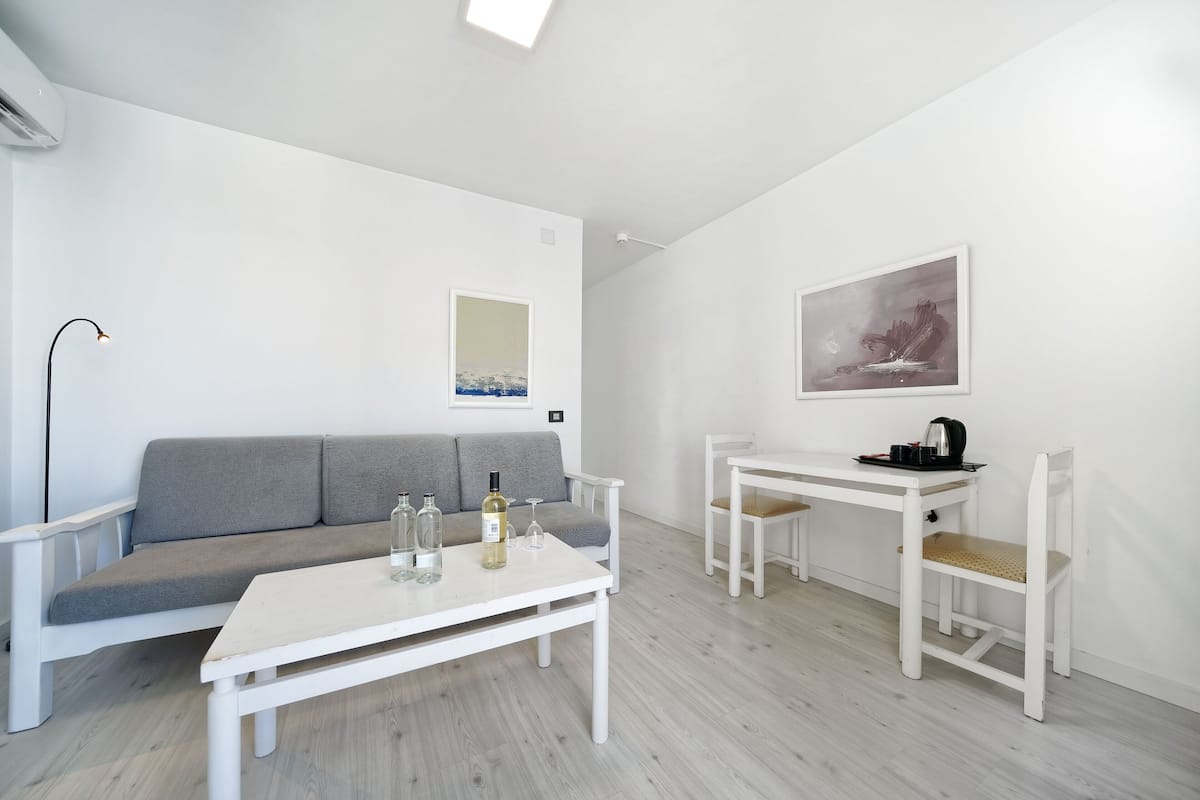 Ein modernes, minimalistisches Wohnzimmer mit einem grauen Sofa, einem weißen Couchtisch mit Flaschen und Gläsern sowie einem weißen Schreibtisch mit Stühlen.