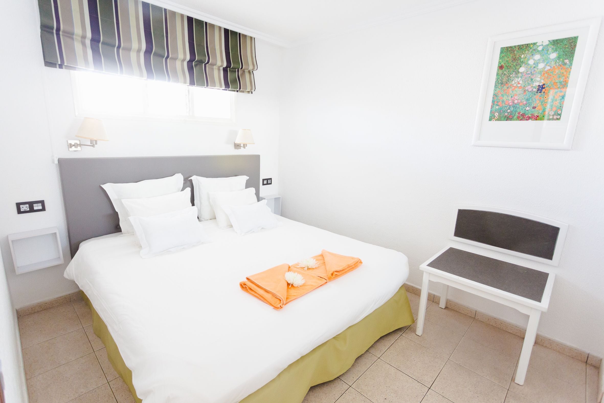 A neatly made bed with white linens, a grey headboard, and a small table with a grey cushion.