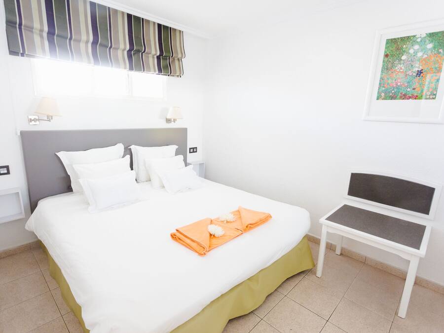 A neatly made bed with white linens, a grey headboard, and a small table with a grey cushion.