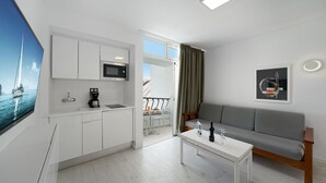 Una cocina moderna con armarios blancos, un microondas y una cafetera. Hay un balcón con vistas al mar. Un sofá gris y una mesa de centro blanca están en la sala de estar.