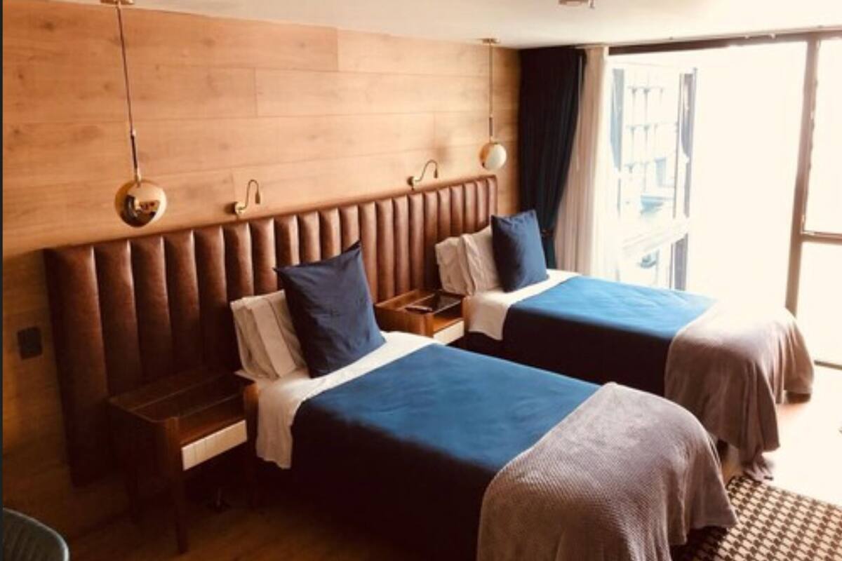A hotel room with two beds, wooden headboards, bedside tables, and a large window with curtains.
