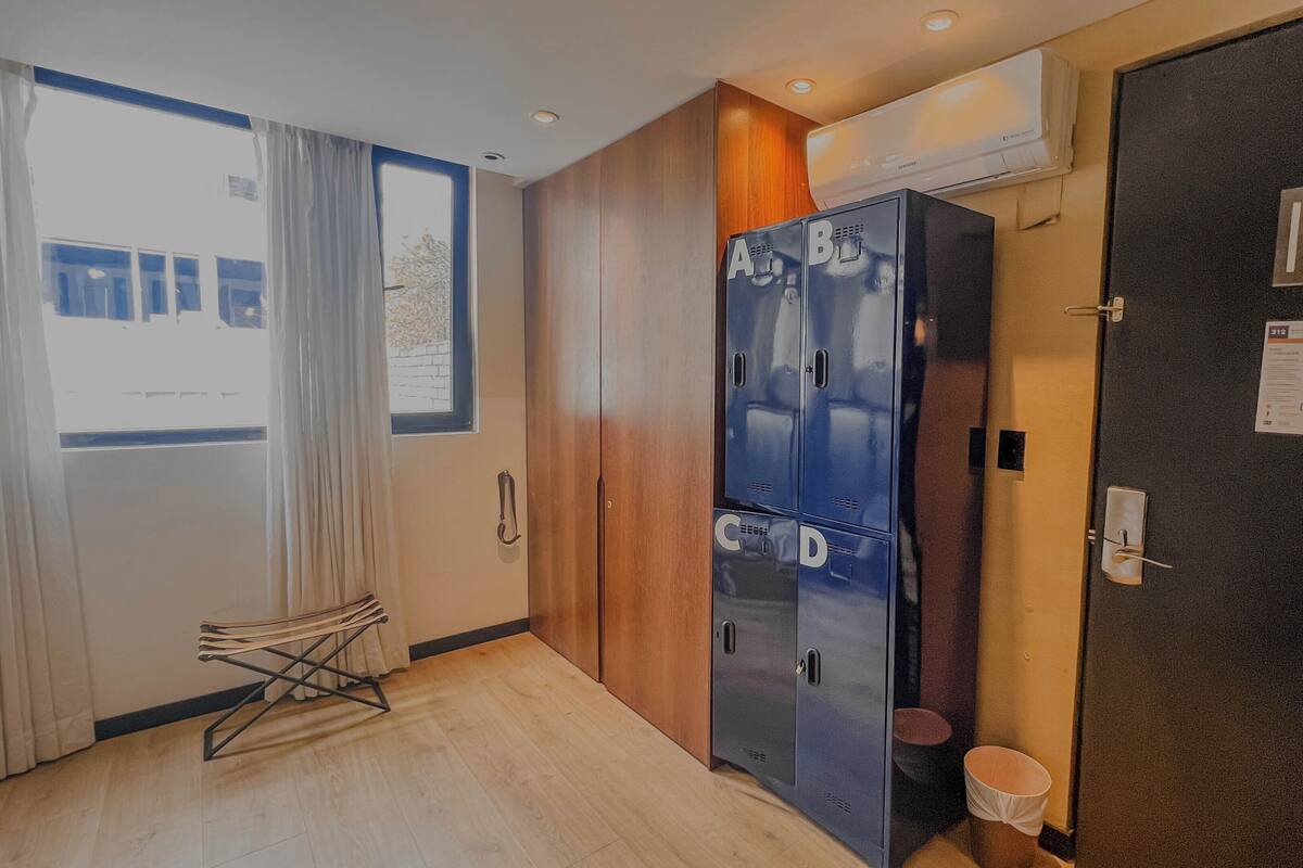 A hotel room with a wooden floor, a window with curtains, a radiator, a wardrobe with multiple compartments, and a door with a light switch and a notice.