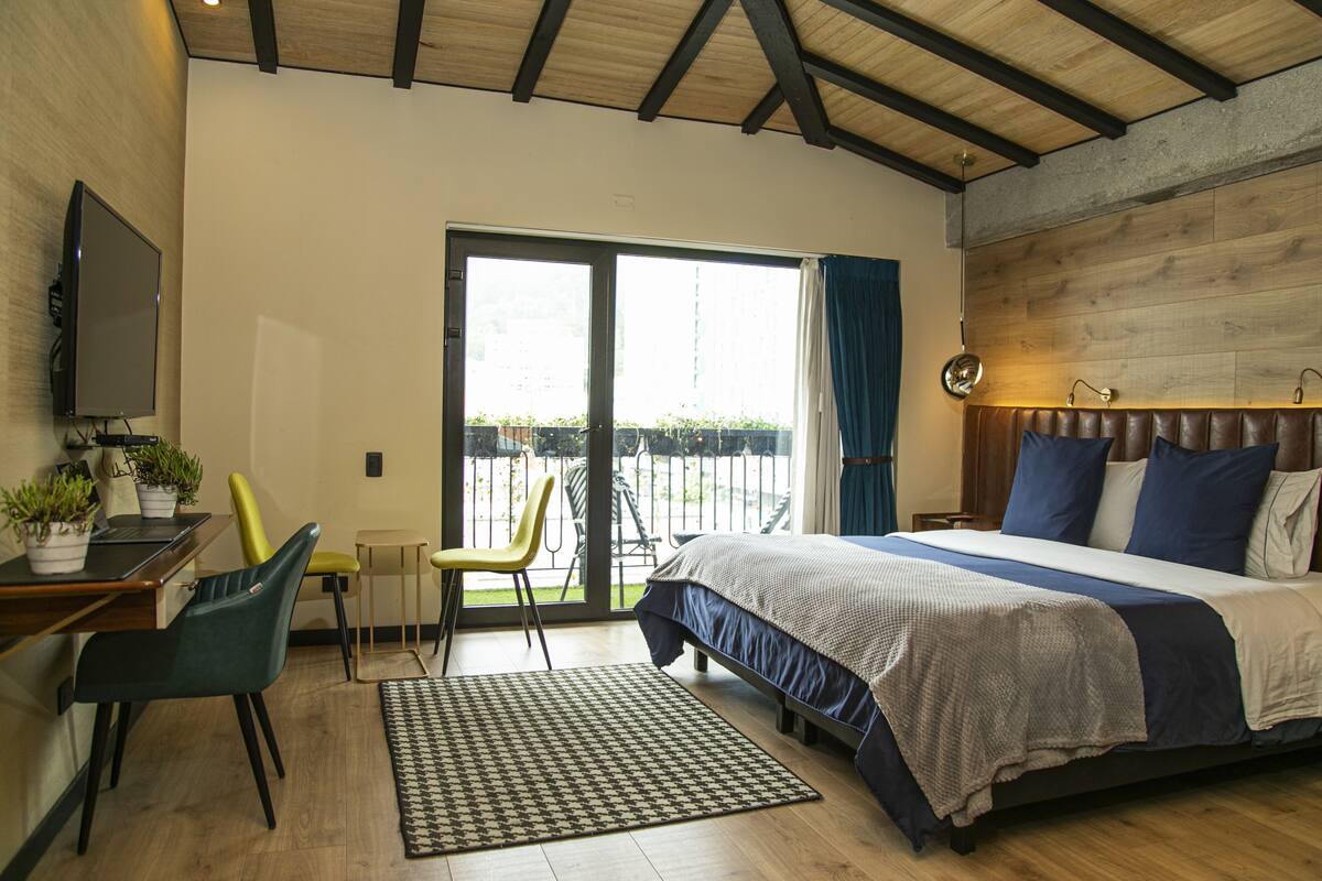 A bedroom with a bed, a desk with a computer, a chair, a window with curtains, and a view of the outdoors.