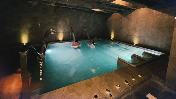 Una piscina tipo spa con dos personas, un muro de piedra y una escalera de metal.
