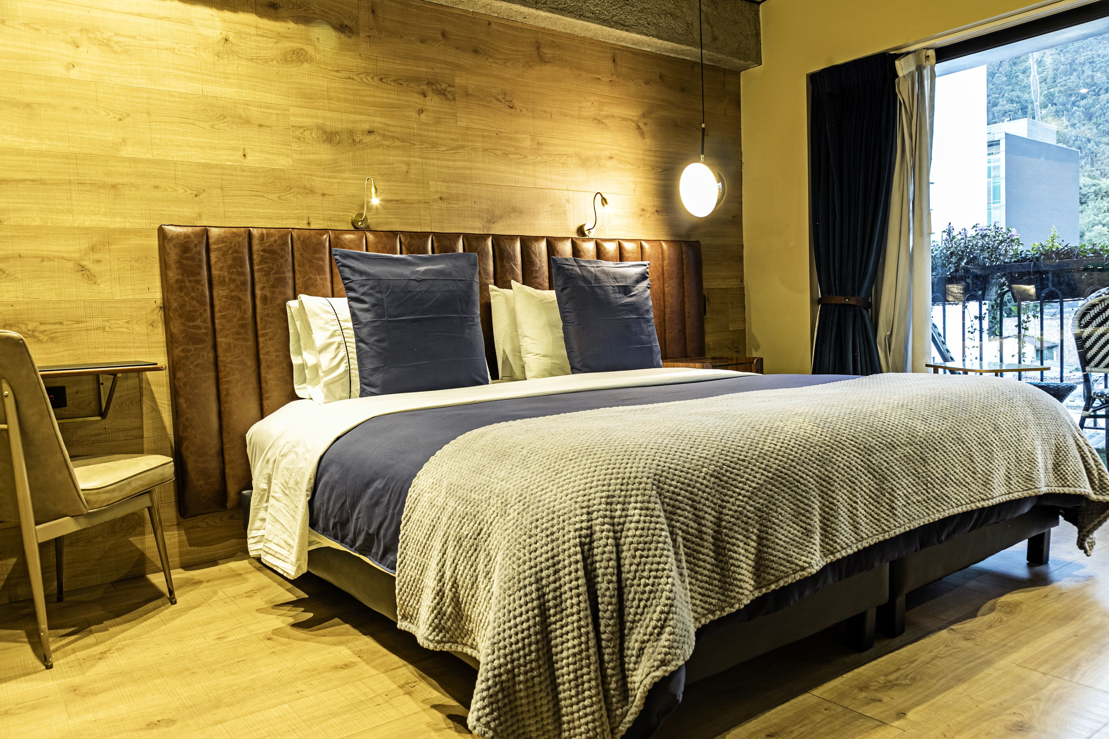 A neatly made bed with a headboard, a bedside chair, and a window with curtains.