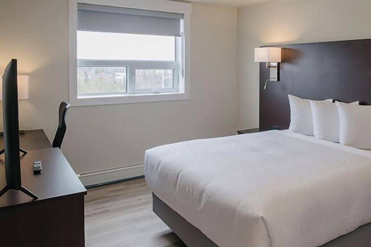 A hotel room with a large bed, a desk with a chair, a window, and a wall-mounted lamp.