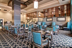 A hotel dining area with multiple tables and chairs, a patterned carpet, and a staircase with wooden railings. - Chateau Nova Yellowknife (Yellowknife)