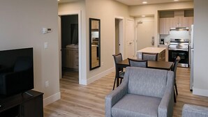 Una sala de estar moderna con un televisor de pantalla plana, un sillón gris y una zona de comedor con una mesa y sillas. Se ve una cocina con electrodomésticos de acero inoxidable al fondo.