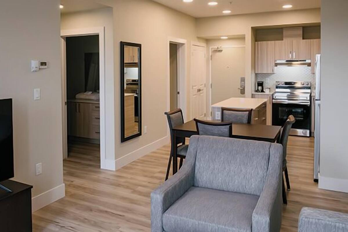 A modern living room with a flat-screen TV, a grey armchair, and a dining area with a table and chairs. A kitchen with stainless steel appliances is visible in the background.