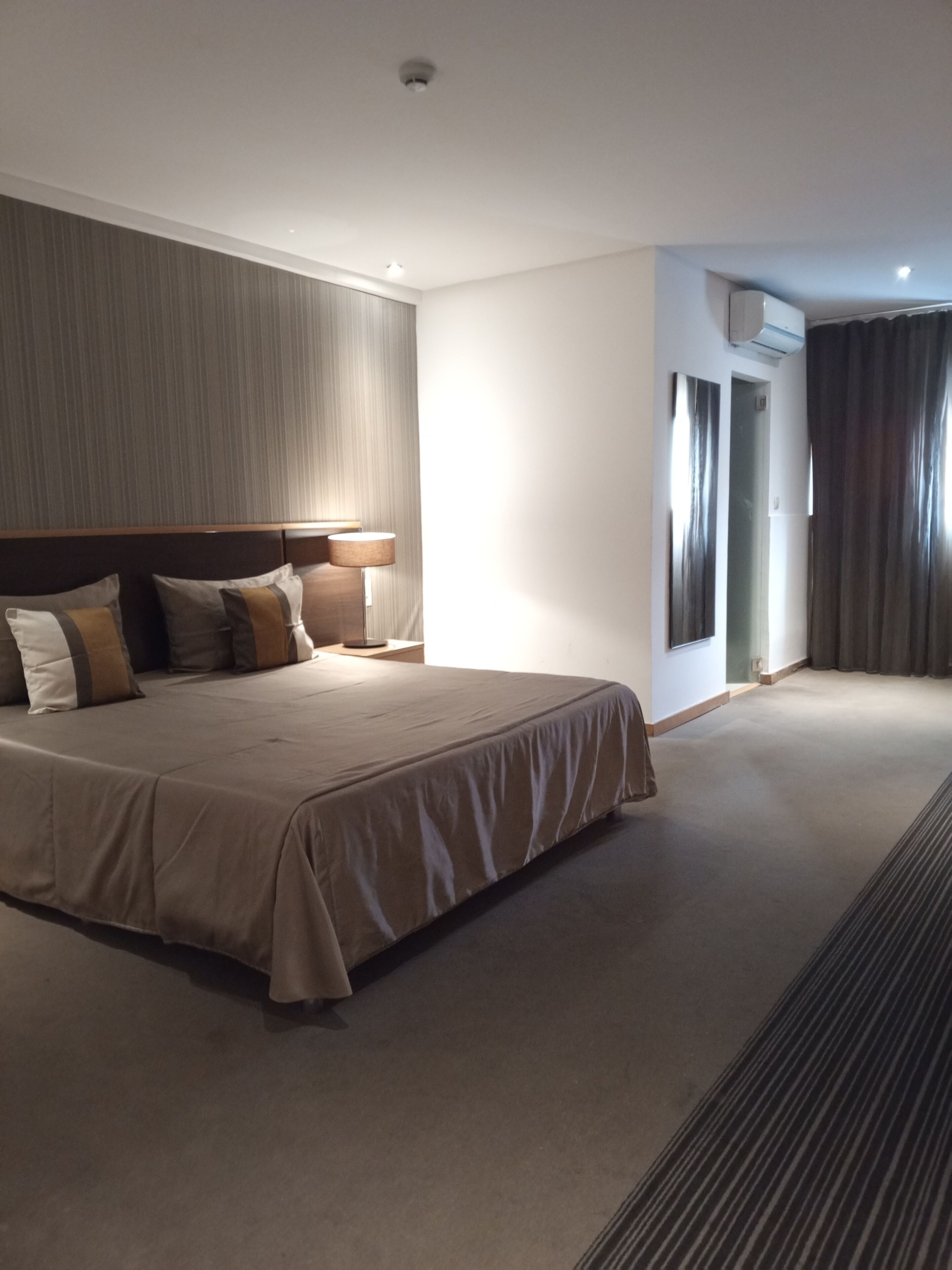 Habitación de hotel con una cama grande, cabecera, mesita de noche y unidad de aire acondicionado.