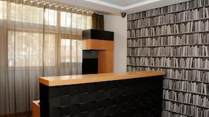 A room with a black and wooden reception desk, a bookshelf filled with books, and a large window with sheer curtains. - Hotel Ritz Capital (Luanda)