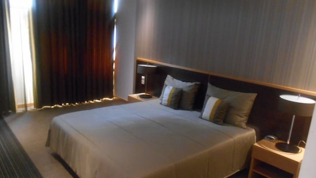 A hotel room with a large bed, a bedside table with a lamp, and a window with curtains. - Hotel Ritz Capital (Luanda)