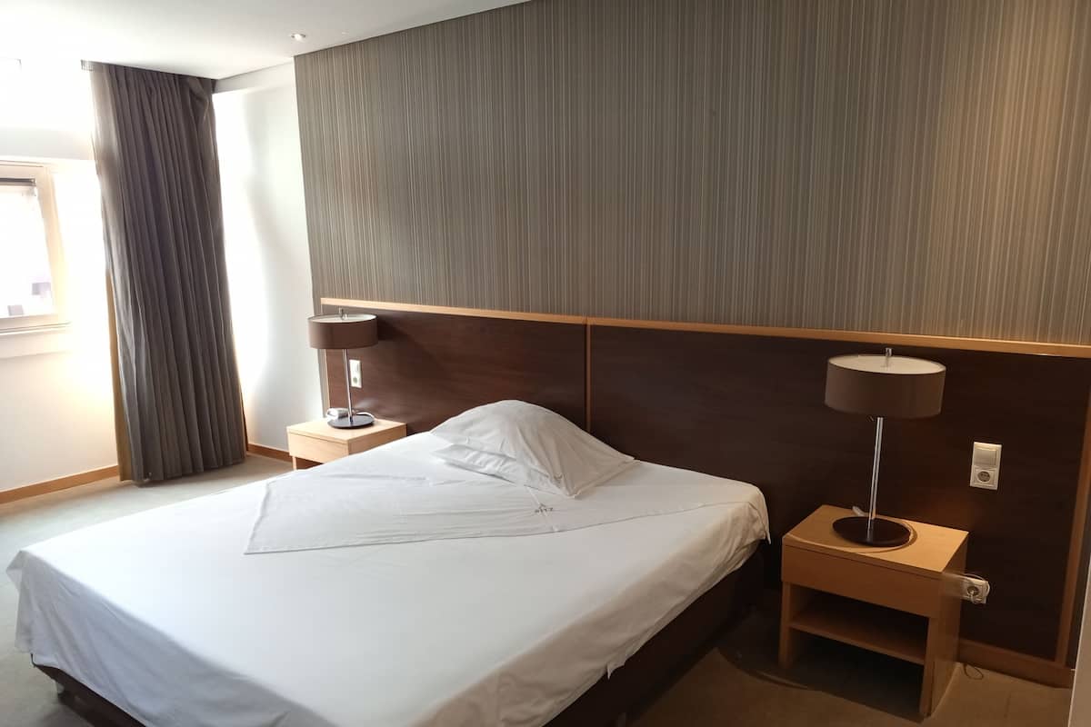 Una cama bien hecha con sábanas blancas, dos mesitas de noche con lámparas, un cabecero de madera y una ventana con cortinas.