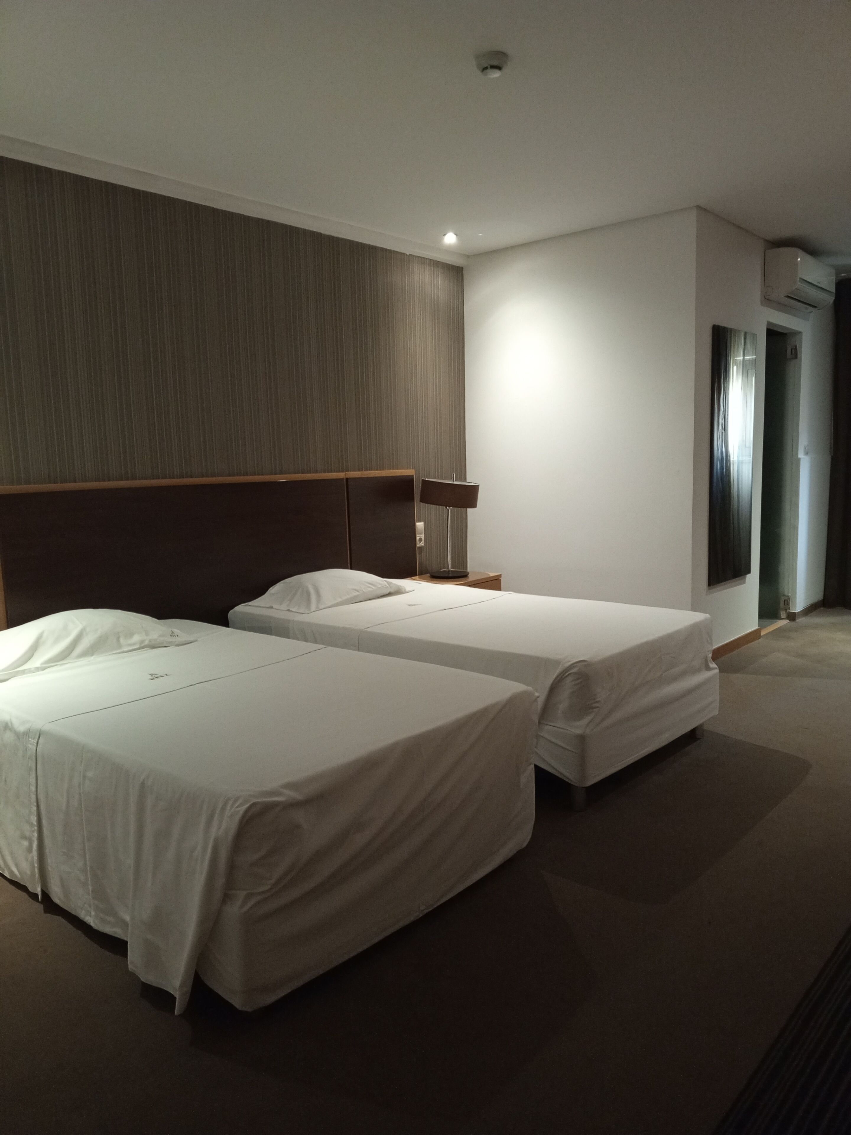 Habitación de hotel con dos camas, cabecera, mesita de noche, lámpara y unidad de aire acondicionado.