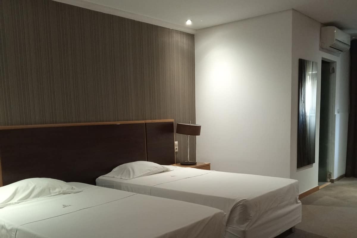 Habitación de hotel con dos camas, cabecera, mesita de noche, lámpara y unidad de aire acondicionado.